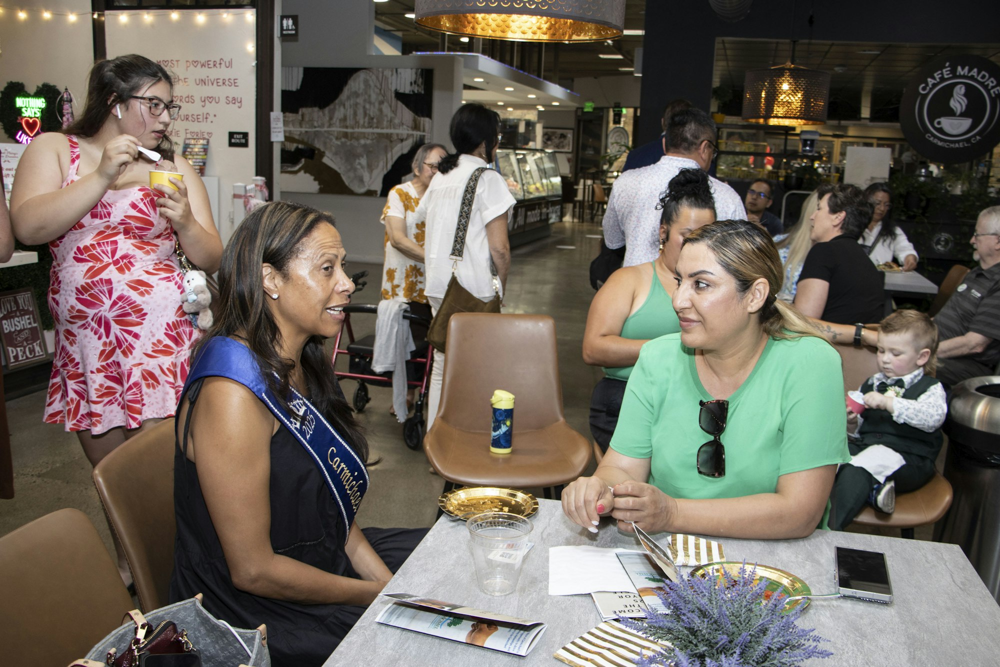 Two women sitting and talking at a table in a busy café or market area. People are eating and chatting around them.