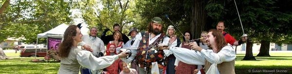 People in historical costumes having a playful sword fight outdoors, with onlookers in the background.