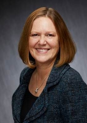 A smiling woman with shoulder-length hair, wearing a blue jacket and a necklace, against a mottled grey background.