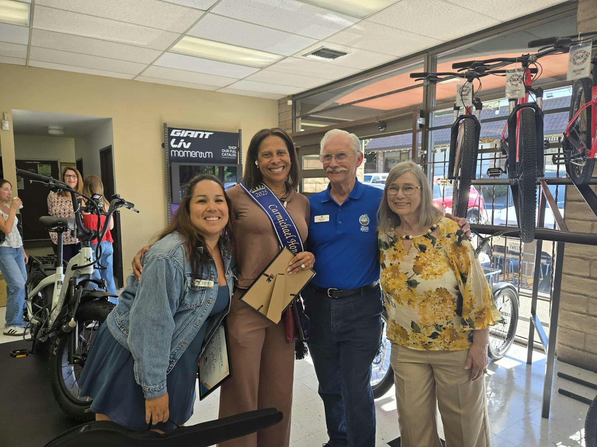 Four people smiling inside a bike shop, one wearing a sash.