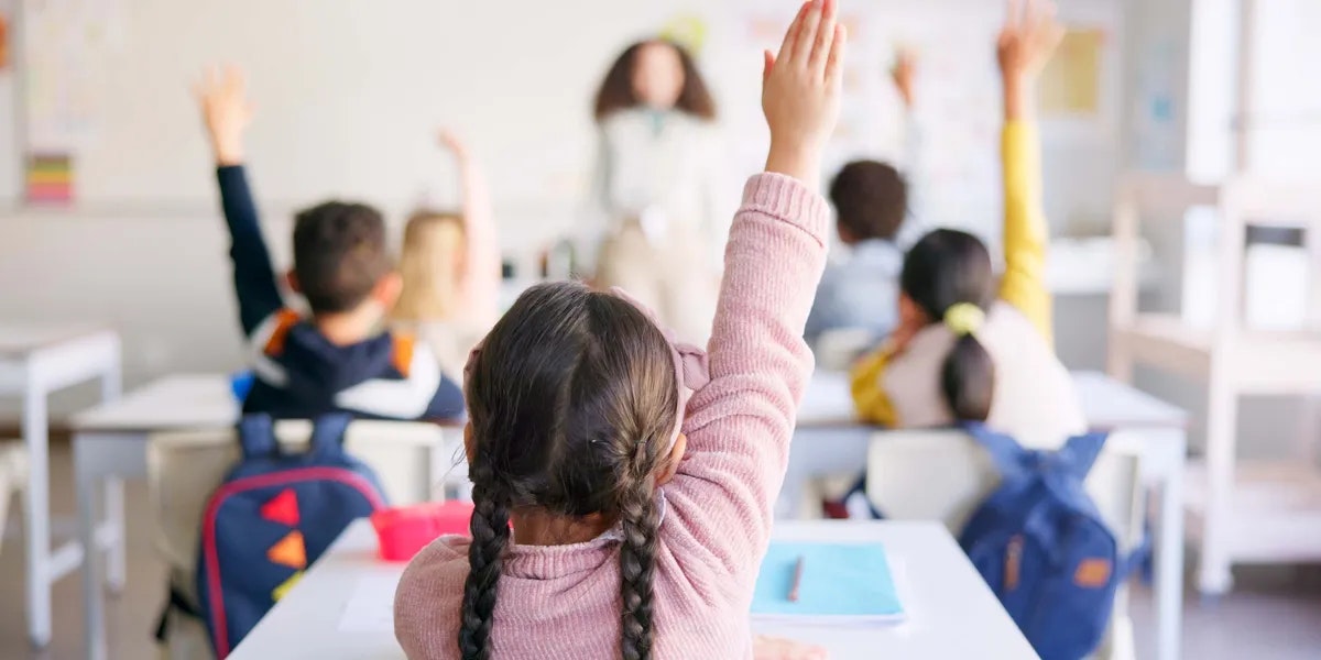 Children in a classroom raising their hands, facing the teacher.