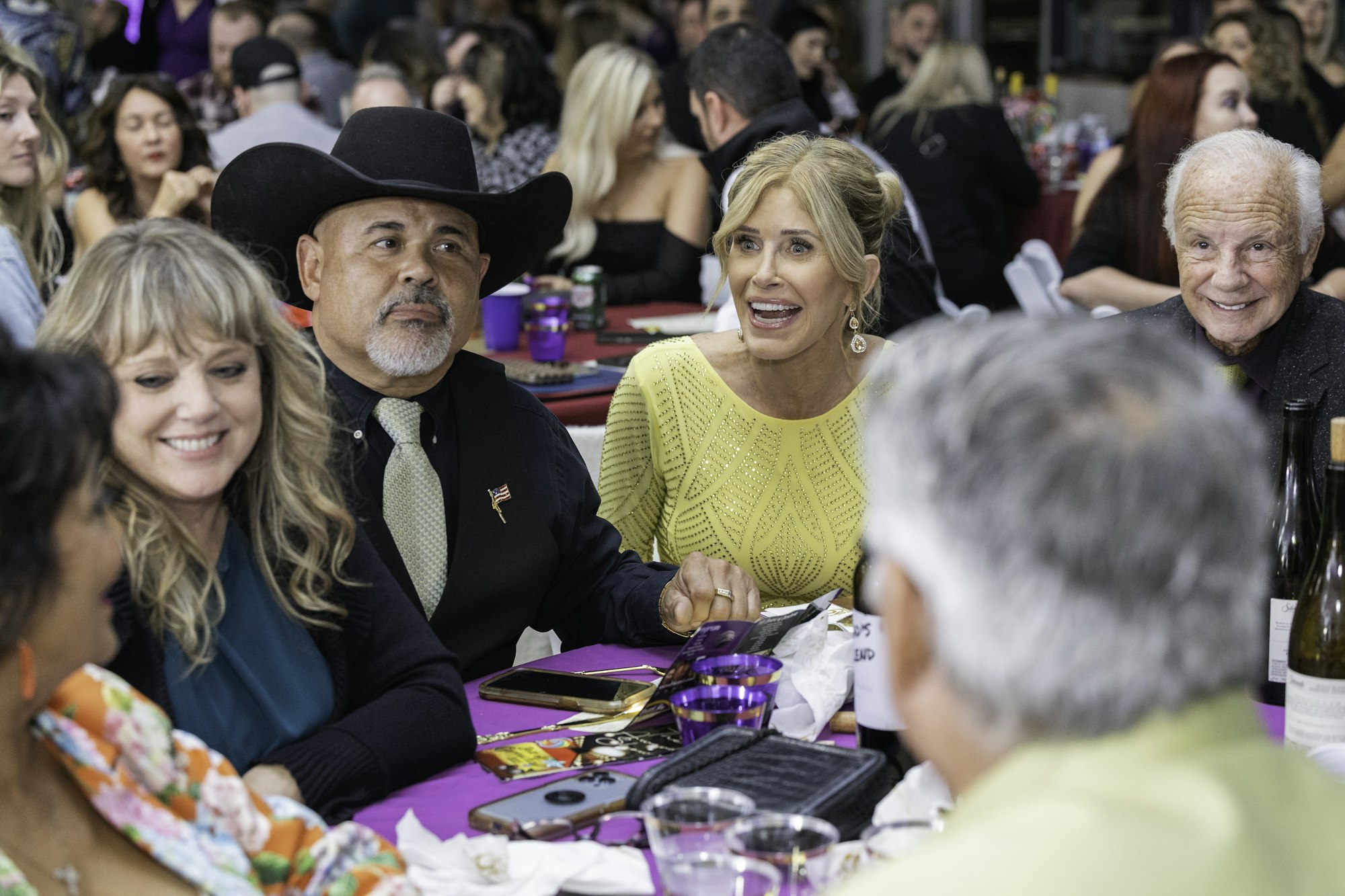 A group of people are seated at a crowded event, smiling and conversing at a table covered with purple tablecloth and drinks.