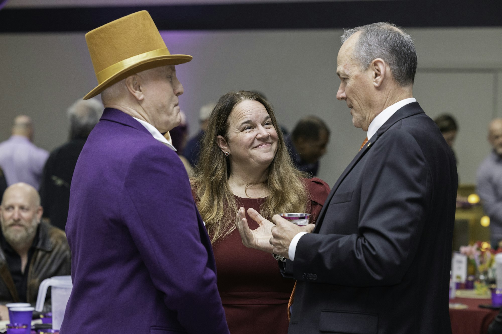 Three people in a conversation at an event. One wears a purple suit and orange hat, another a black suit, and the third a red dress.