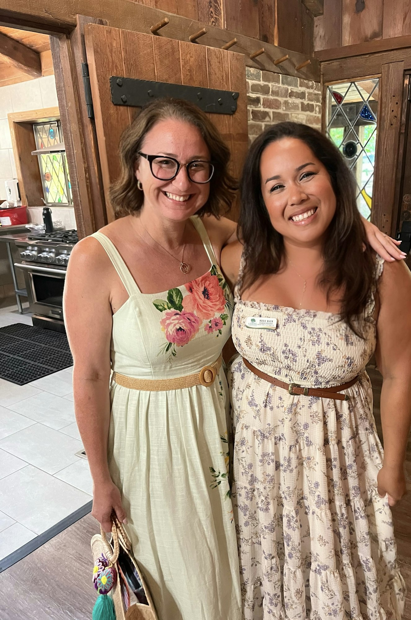 Two smiling women in floral dresses stand together in a rustic room.