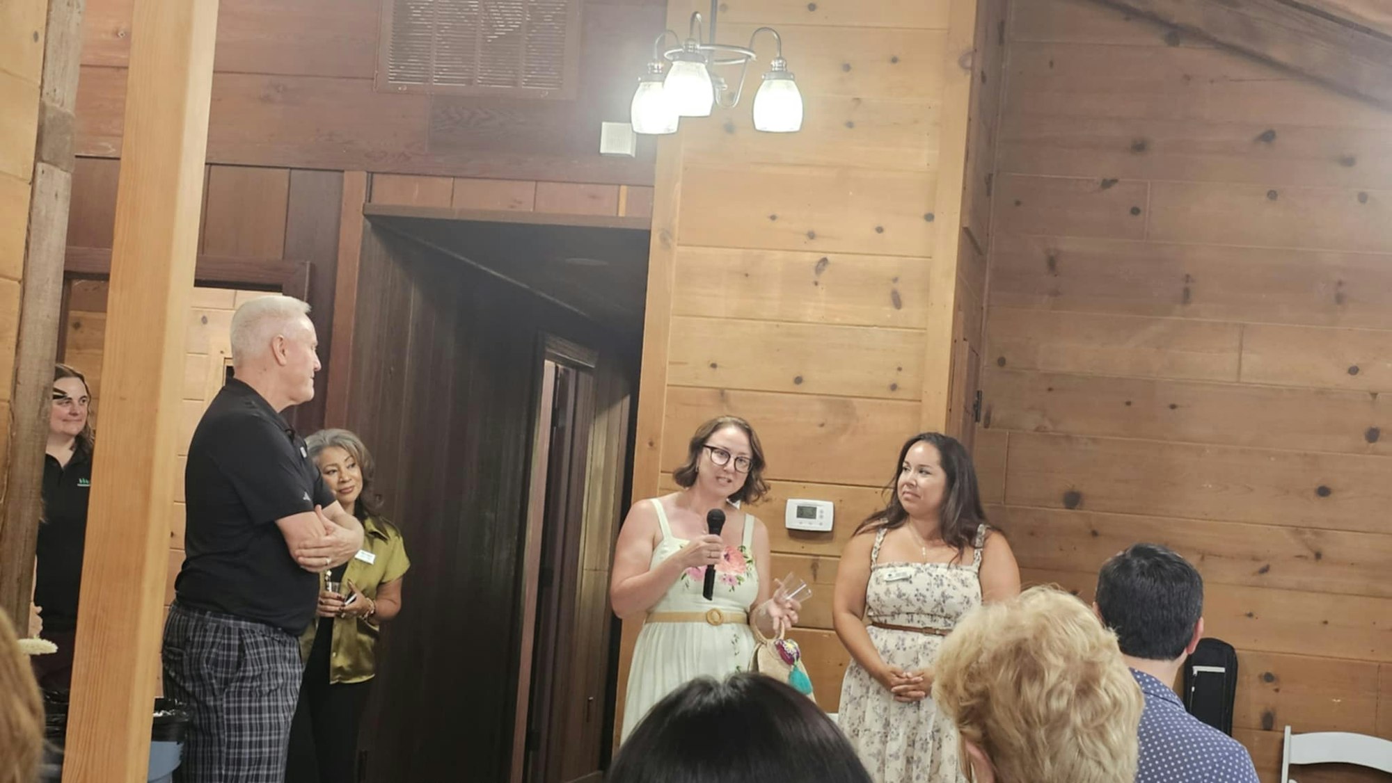 A woman speaks into a microphone while others listen in a wooden room setting.