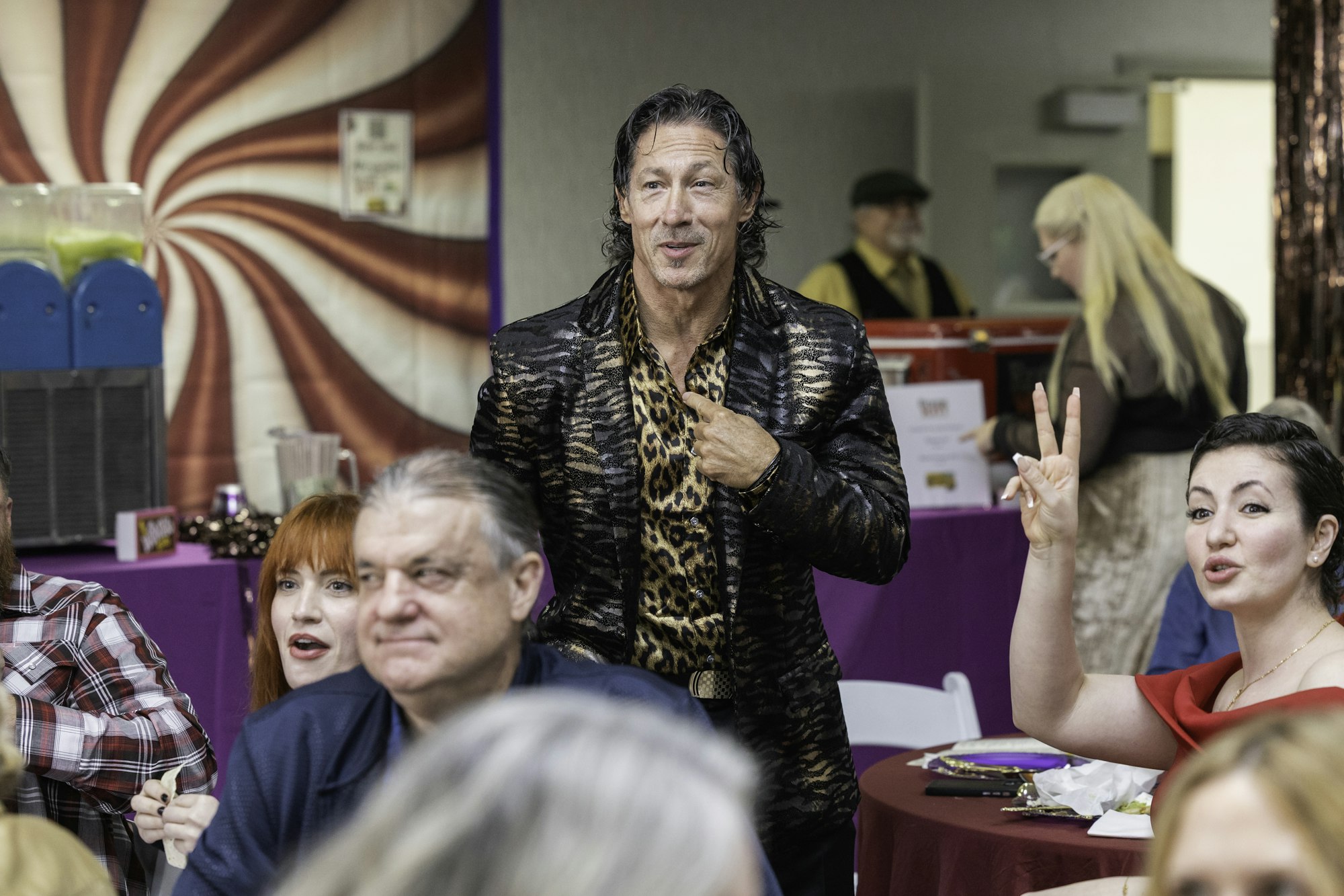 A man in a leopard-print shirt stands in a crowd; a woman gestures peace sign, colorful background.