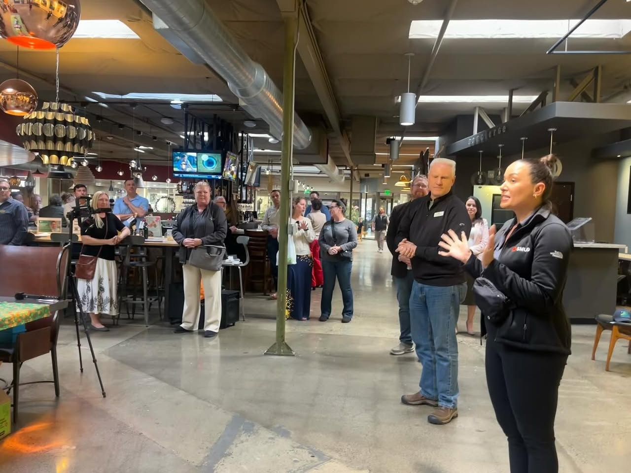 A group of people in a modern indoor space, listening to a speaker gesturing as she addresses them.