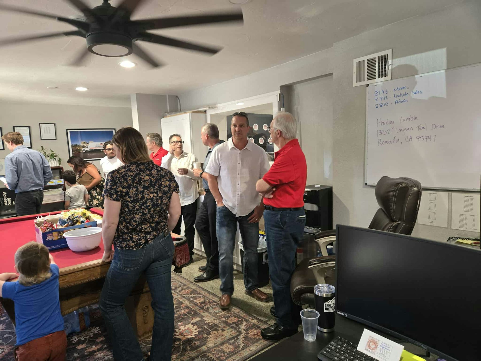 A group of people socializing in a room with snacks, a pool table, and a whiteboard displaying an address.