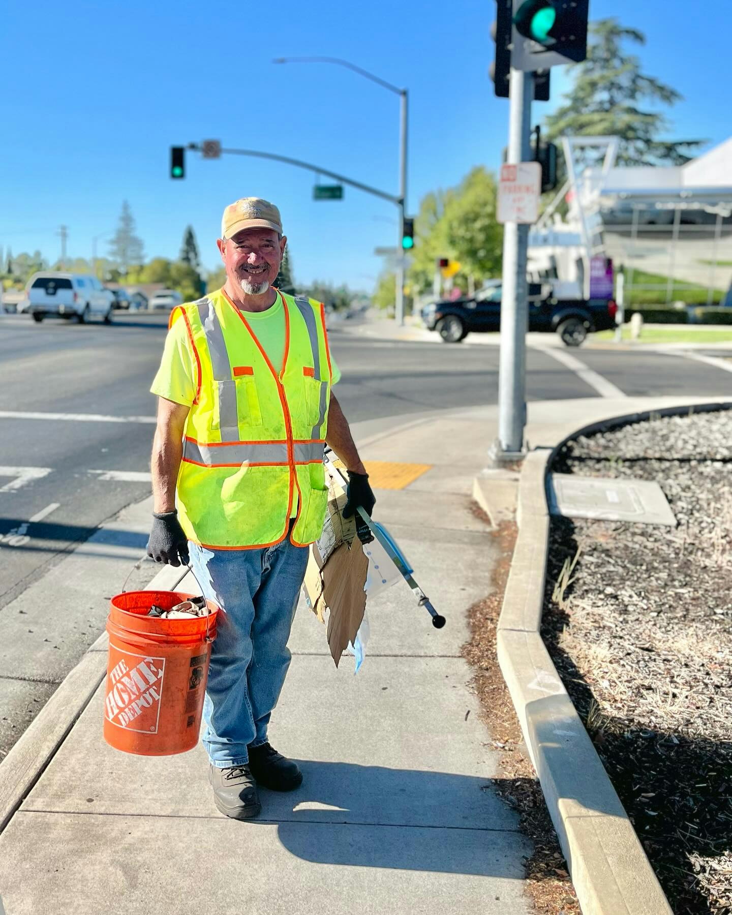 A person in a high-visibility vest stands at a crosswalk holding a bucket and picker tool, near a traffic light.