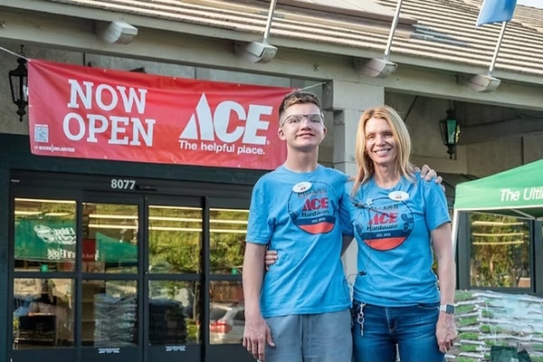 A woman and a young man pose in front of an Ace Hardware store with a "Now Open" banner. Both are wearing matching blue shirts.