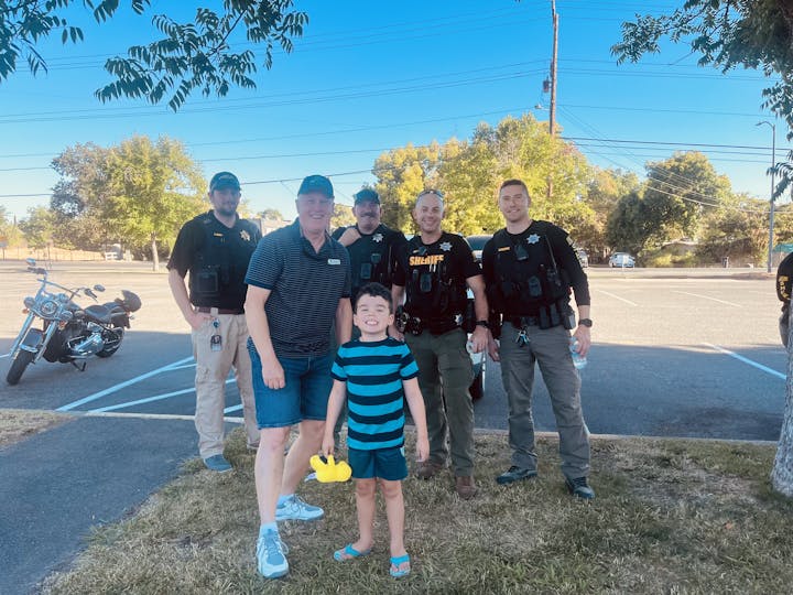 A child and adult pose with four law enforcement officers in a parking lot; a motorcycle is in the background.