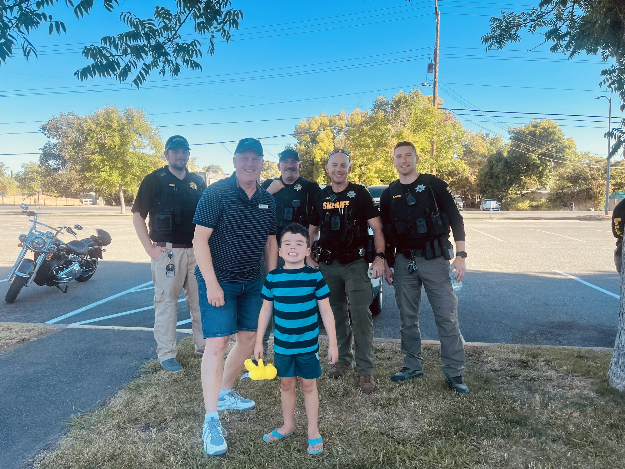 A child and adult pose with four law enforcement officers in a parking lot; a motorcycle is in the background.