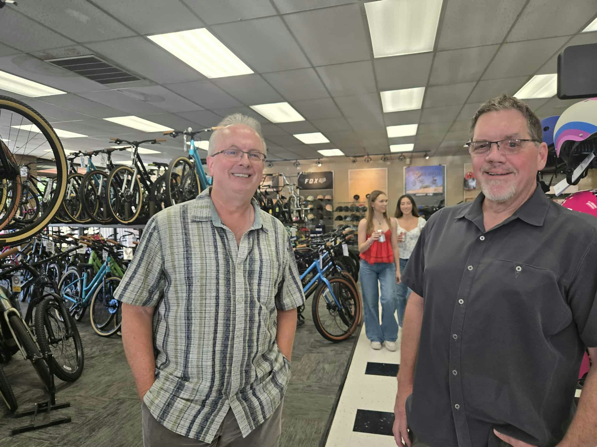 Two men in a bike shop with bicycles, helmets, and two women in the background.