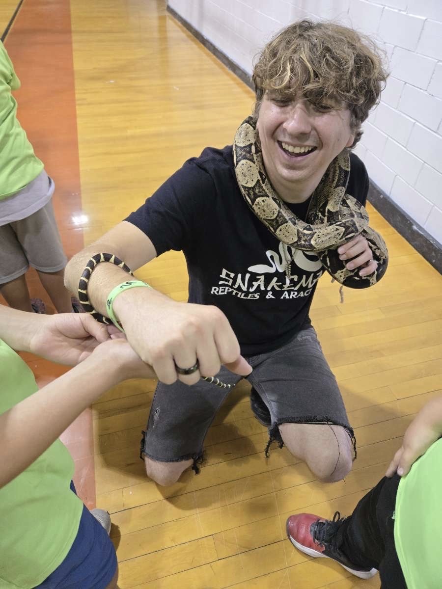 A person with a snake around their neck, smiling, interacting with others in a gym or similar setting.