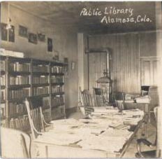 Old library interior with shelves, wooden chairs, tables, books, and a stove. The sign reads "Public Library Alamosa, Colo."