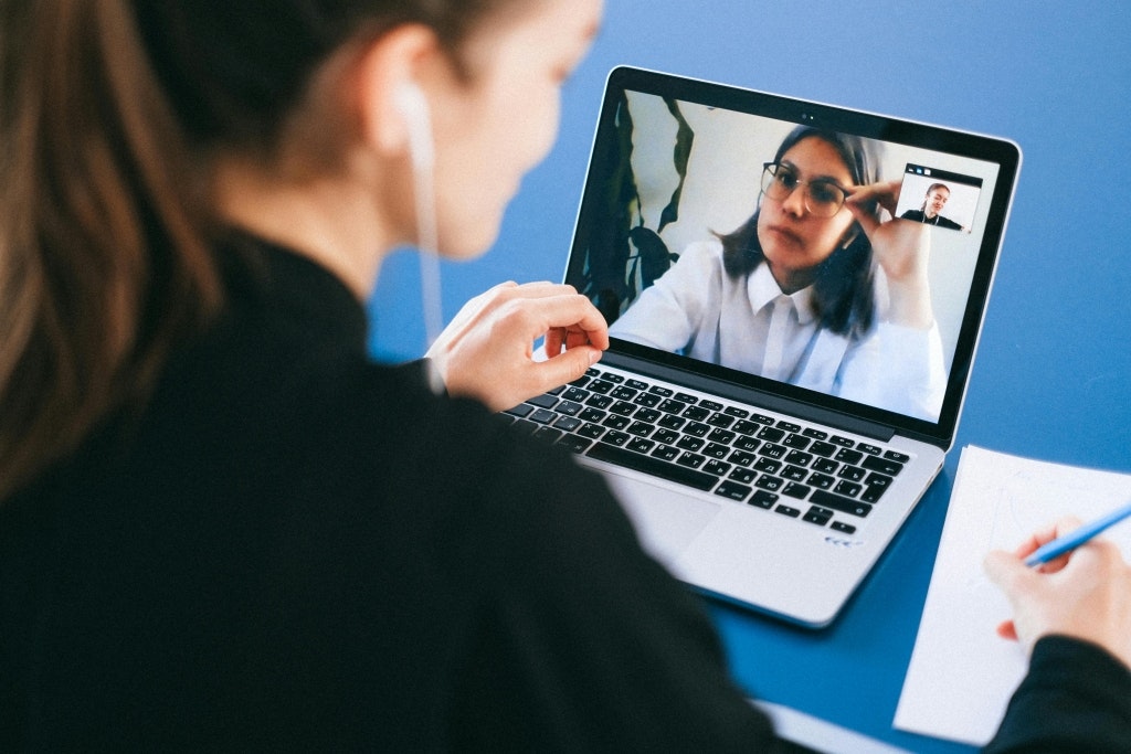 A person having a video call on a laptop, taking notes, and wearing earphones.