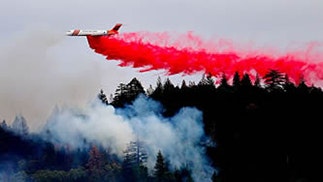 An airplane dropping red fire retardant over a forested area with smoke rising, likely combating a wildfire.