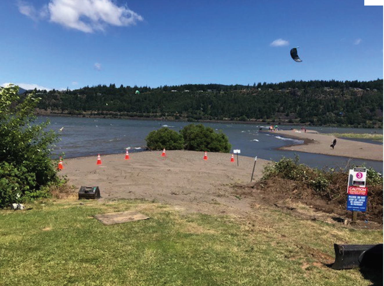 A riverside with people, kitesurfing in the distance, signs, and traffic cones on sandy ground.