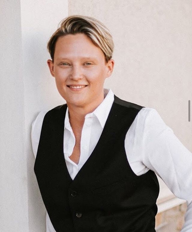 A person smiling, wearing a white shirt and black vest, leaning against a wall.