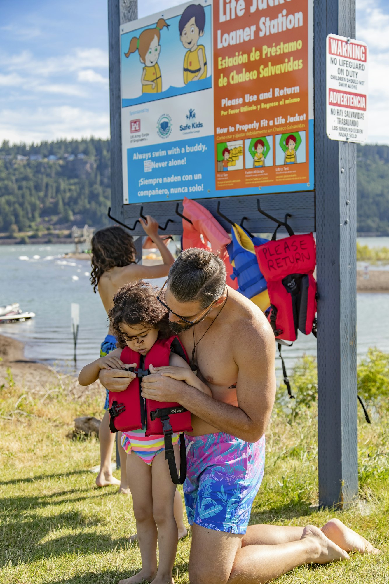 A parent helps a child wear a life jacket at a loaner station by a lake, with safety signs and other children in the background.