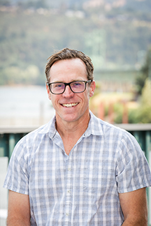 A smiling person wearing glasses and a checkered shirt, with a blurred background that suggests an outdoor setting.