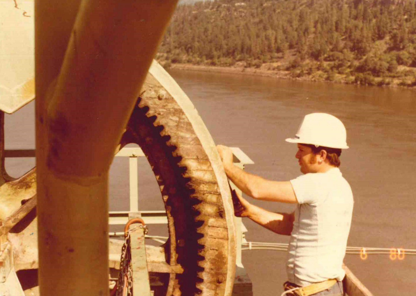 A worker in a hard hat adjusts a large gear by a body of water. Vintage industrial setting.