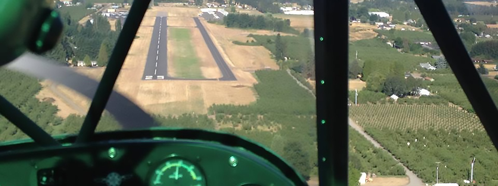 View from an airplane cockpit approaching a runway, with fields and trees surrounding the airstrip.