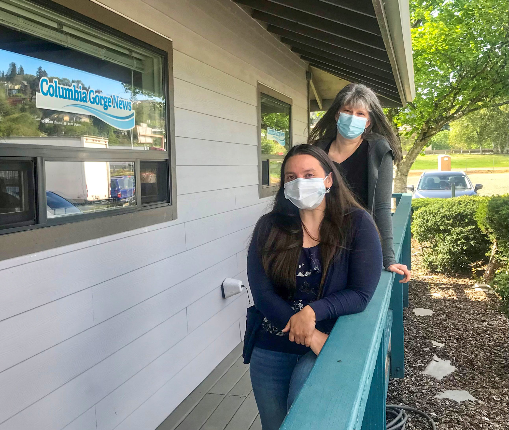 Two people wearing masks leaning on a railing outside a building with a "Columbia Gorge News" window sign.
