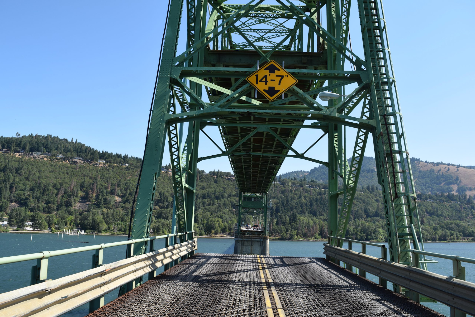 A metal truss bridge with a clear height restriction sign over a scenic waterway flanked by forests and hills.