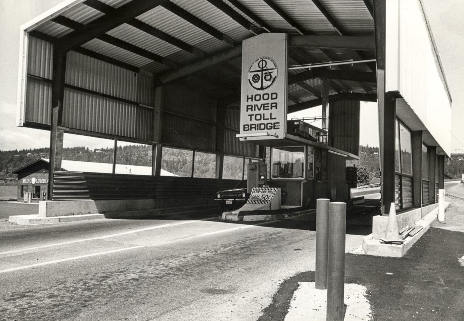 Black and white photo of the Hood River Toll Bridge booth with signs and a car approaching.