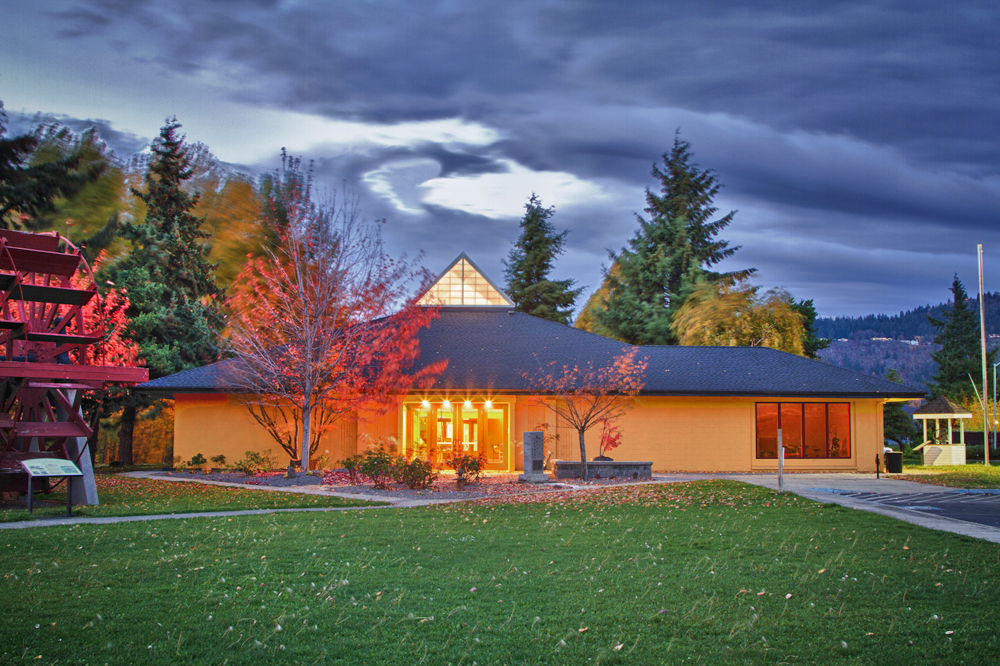 A lit building at dusk with a green lawn and trees, under a dramatic sky.