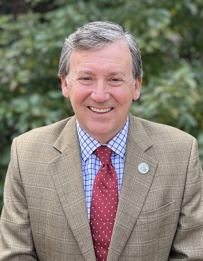 A smiling person in a checkered jacket, blue checkered shirt, red dotted tie, with foliage background.