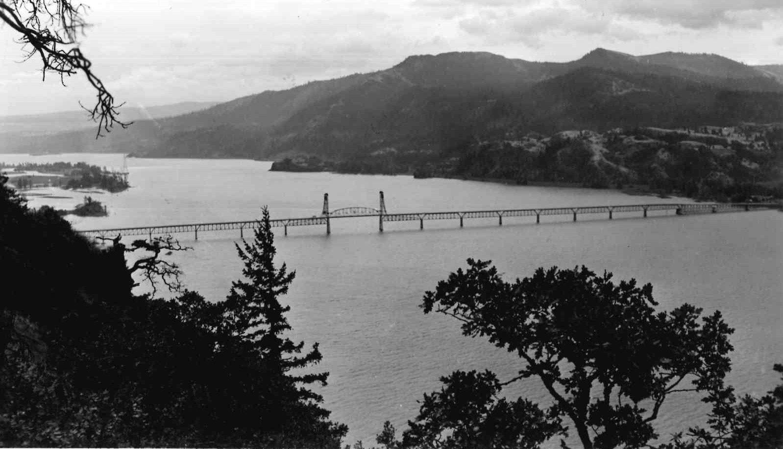 Black and white photo of a bridge spanning a river with forested mountains in the background.