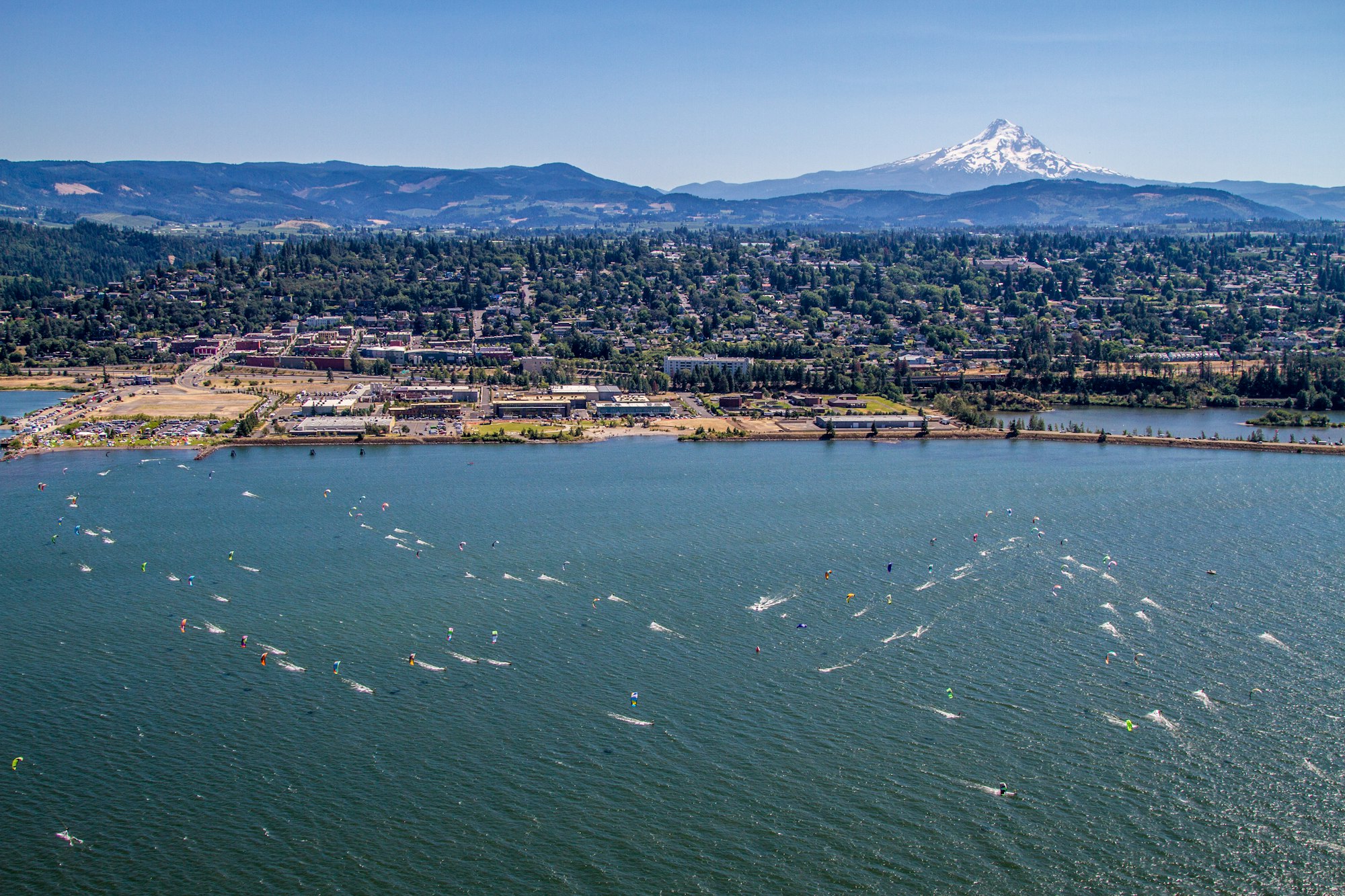 Aerial view of kiteboarders on a river, a town with greenery, and a snow-capped mountain in the background.