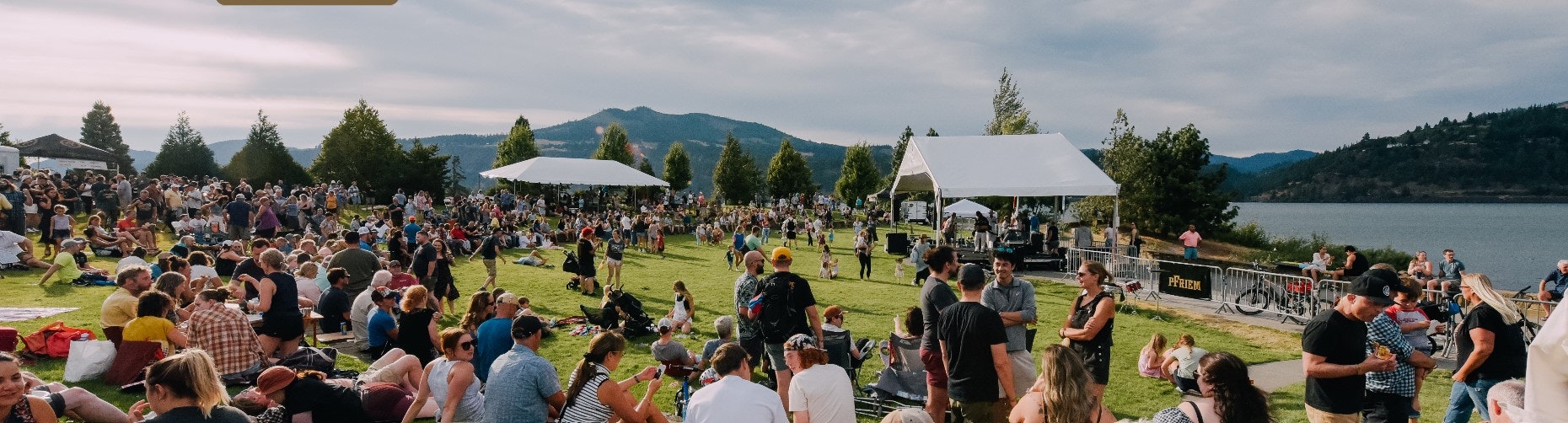 Crowd at an outdoor event by a lake, with tents, trees, and mountains in the background.