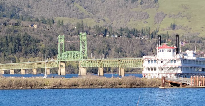 A scenic view featuring a green bridge, a steamboat docked by the river, and hills in the background. Nature and architecture blend beautifully.