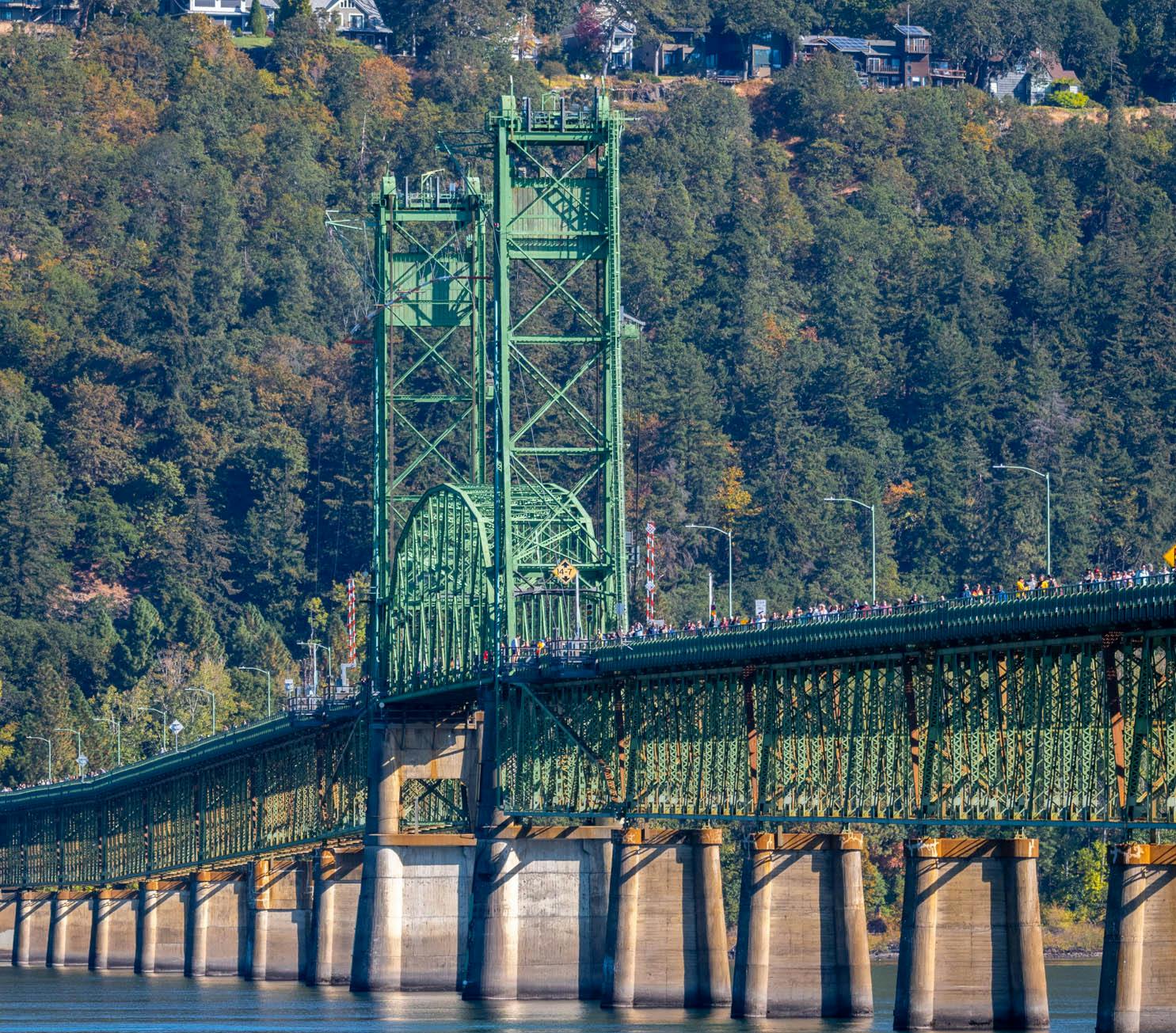 Green truss bridge over a river, surrounded by forested hills and houses.