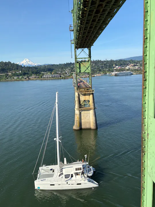 A white sailboat is anchored beneath a green bridge, with a mountain and a scenic landscape in the background.