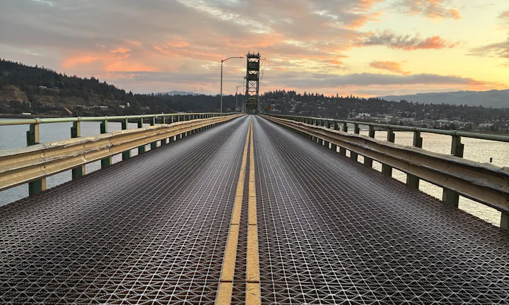A view of a bridge with a metal grate surface, leading toward a sunset with hills and a town in the background.
