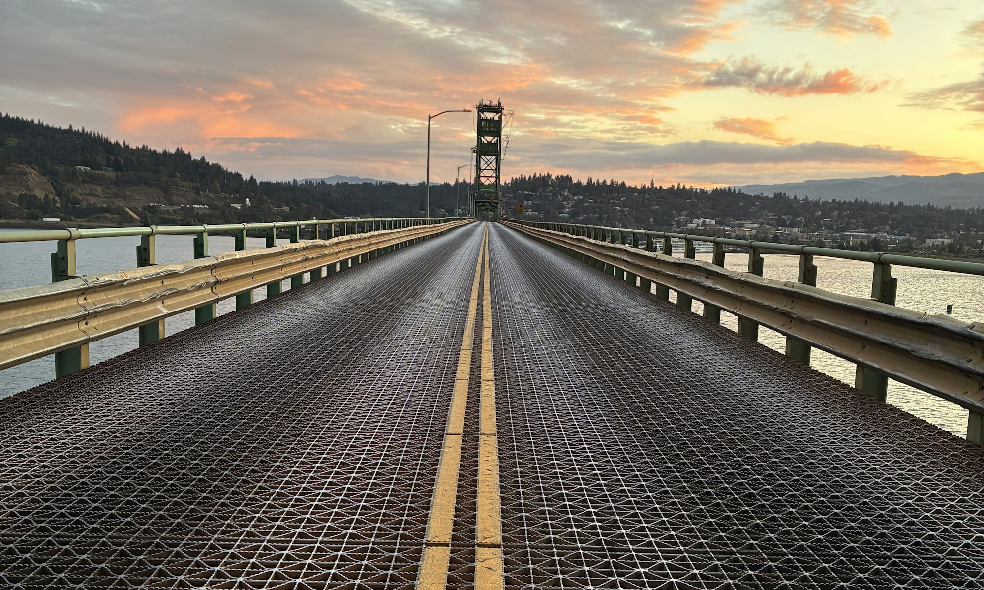 A view of a bridge with a metal grate surface, leading toward a sunset with hills and a town in the background.