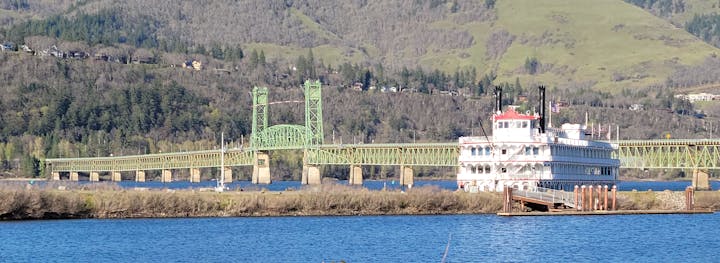 A scenic view featuring a vibrant green bridge and a riverboat on calm waters, surrounded by lush, hilly landscapes.