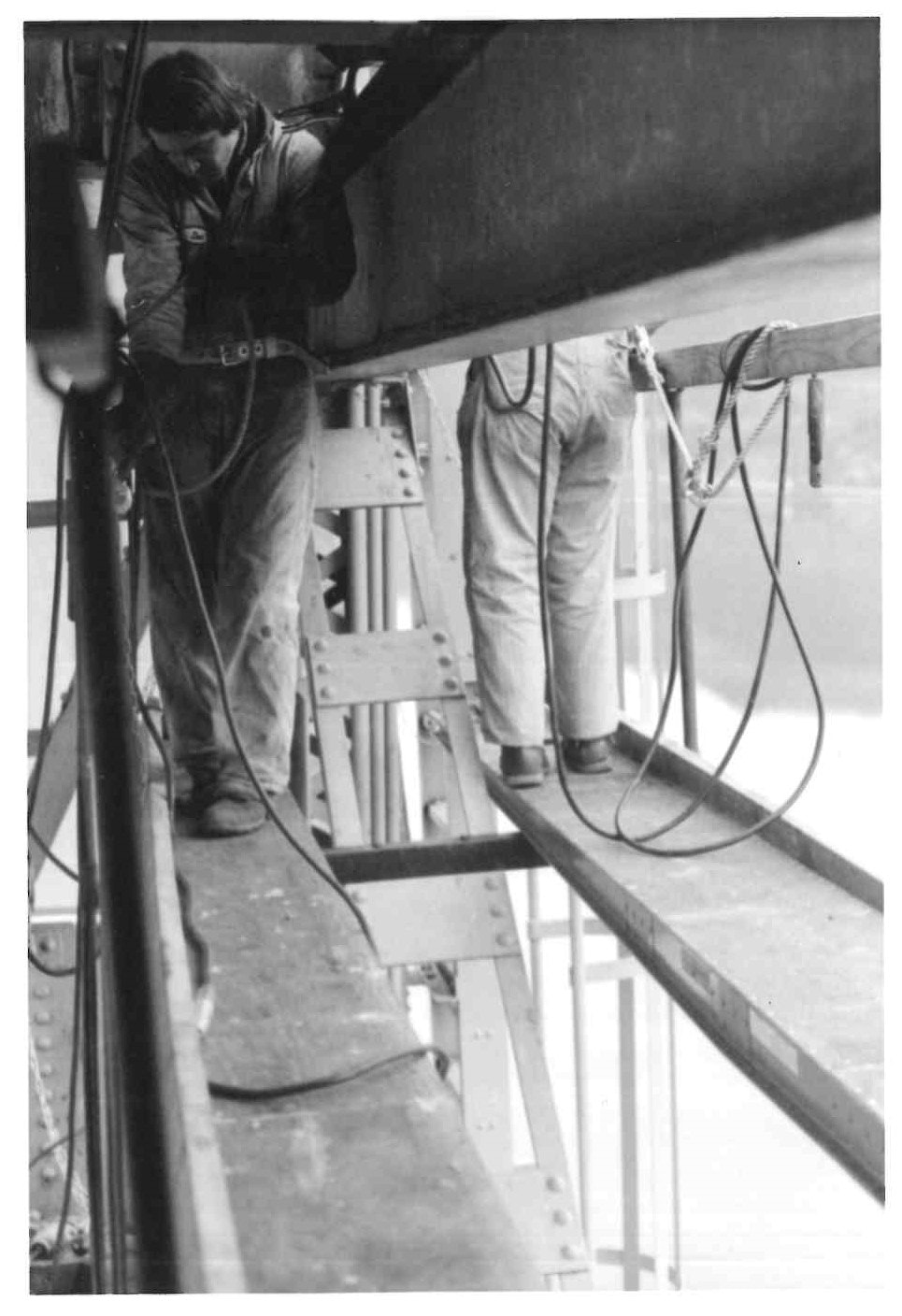 Two workers on a high metal structure, with safety harnesses, possibly engaged in construction or maintenance.