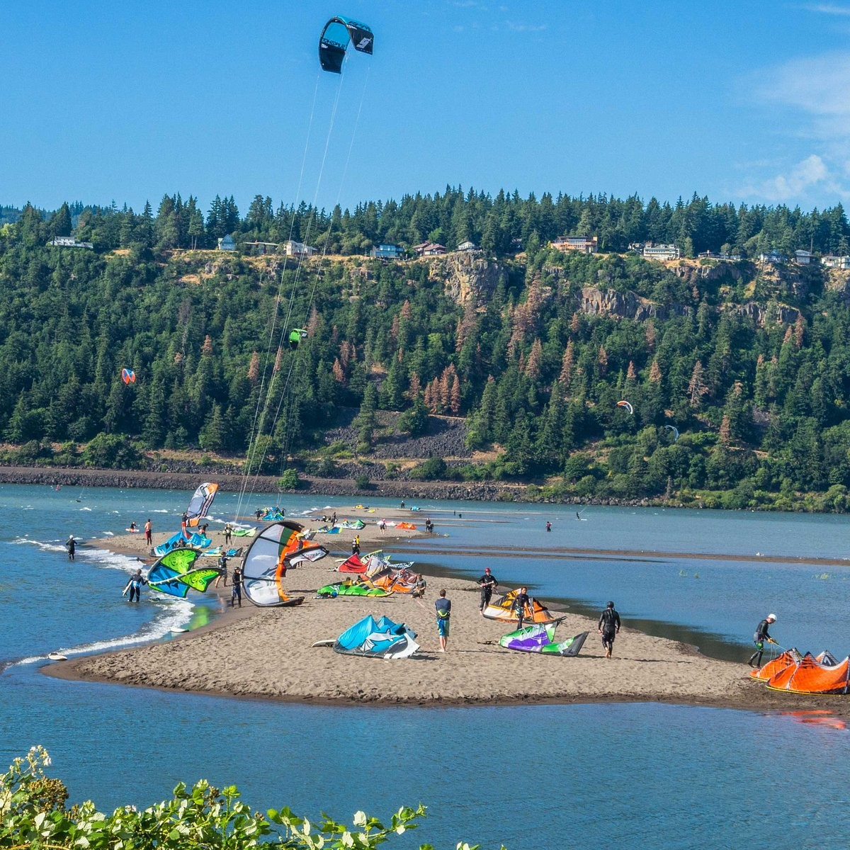 Kiteboarders preparing and launching on a sandy riverbank with a forested hill in the background.