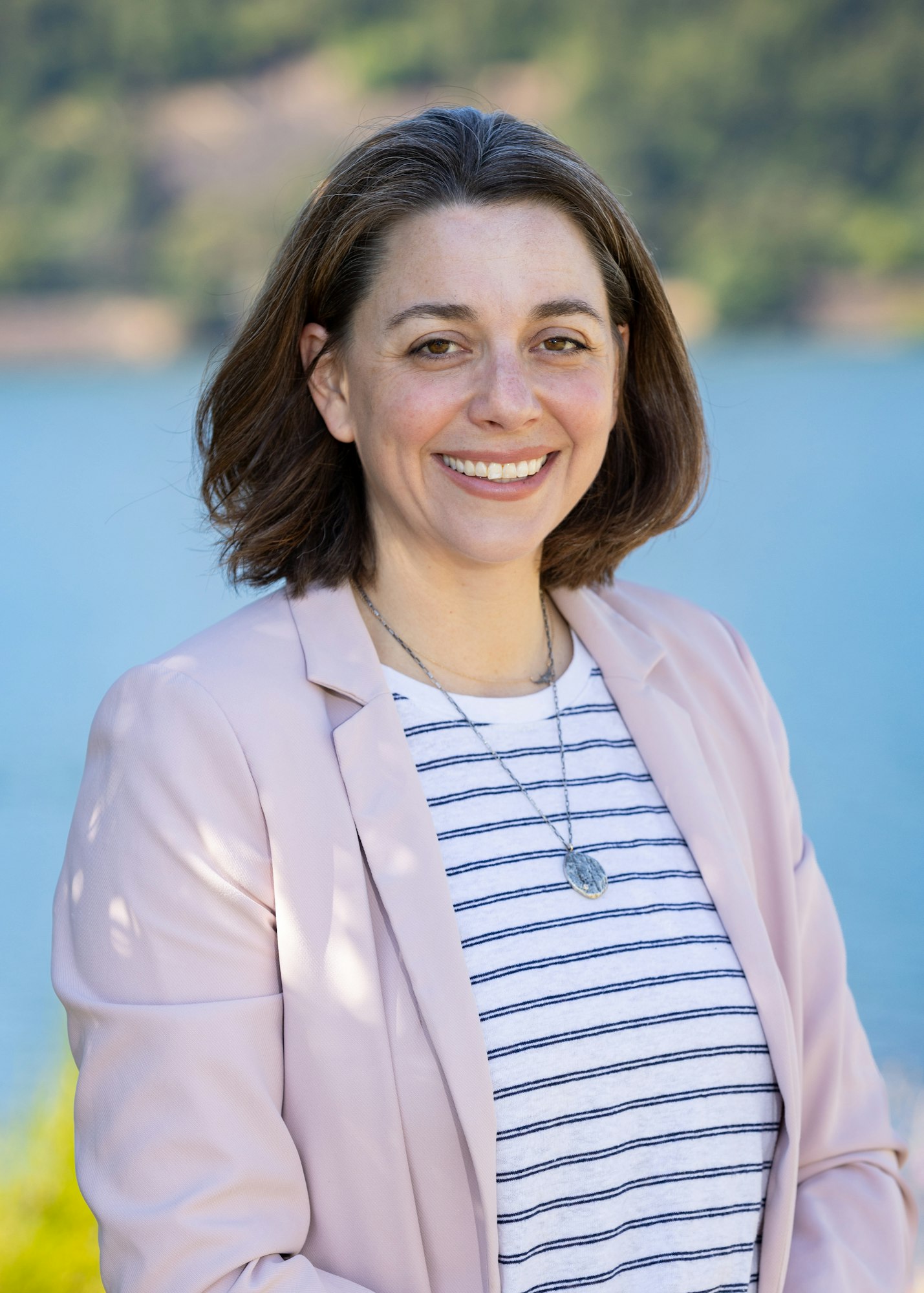 A person smiling at the camera, wearing a striped shirt and pink blazer, standing outdoors near water.