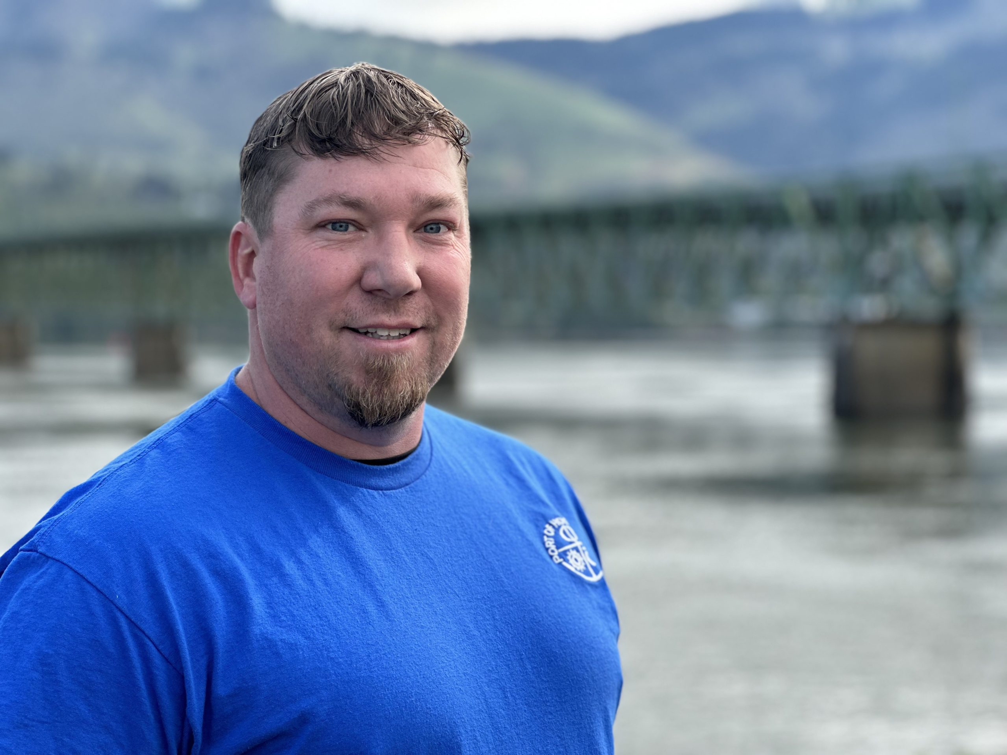 A smiling man in a blue shirt with a bridge and hills in the background.