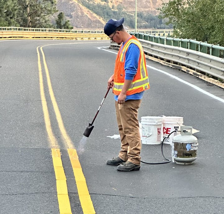 A worker with safety vest painting road lines next to buckets and equipment.