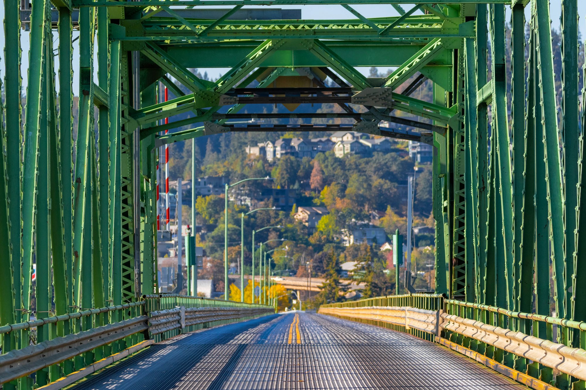 A green metal bridge with a road leading to a hilly area with houses and trees.