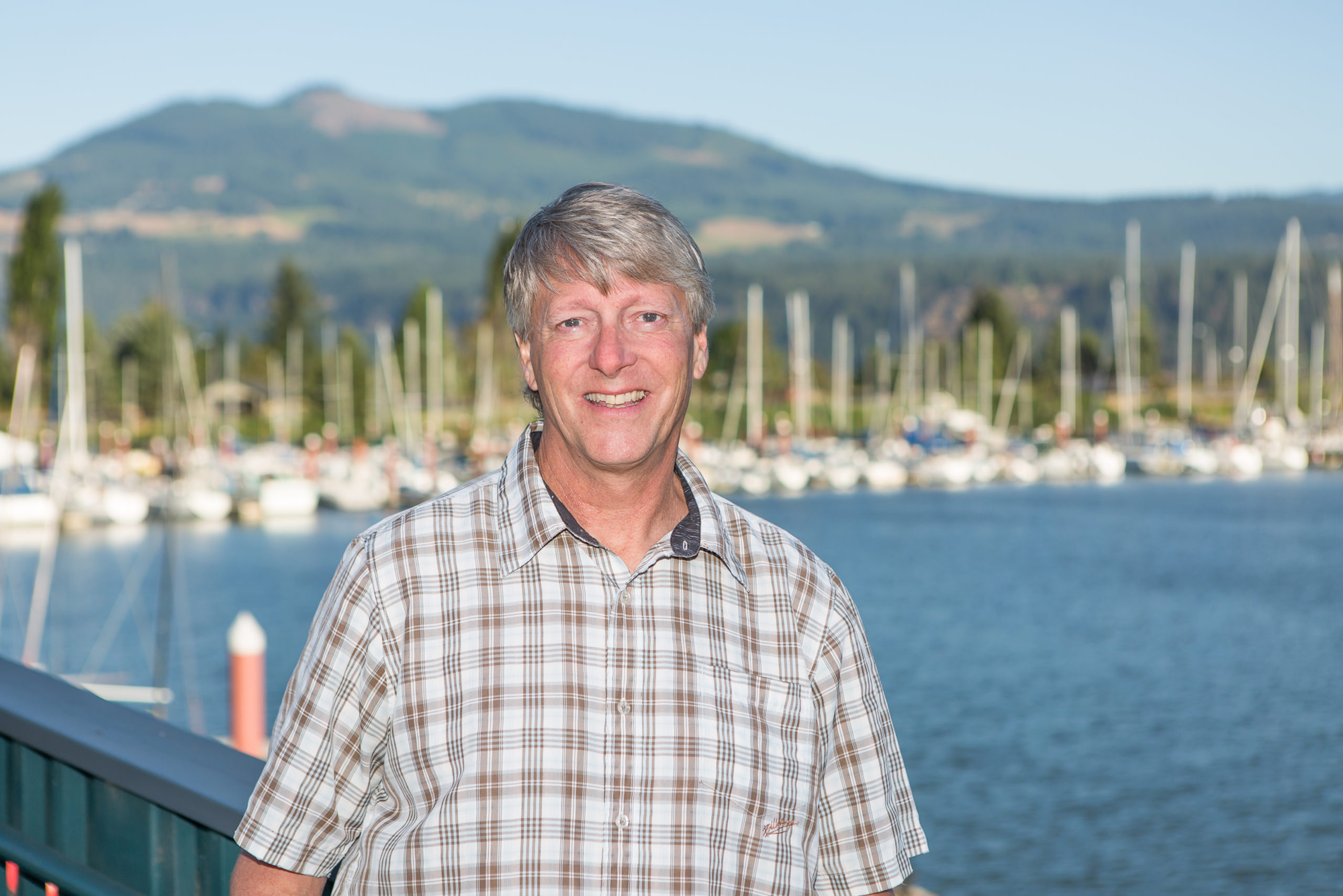 Man smiling at a marina with boats and a mountain in the background.