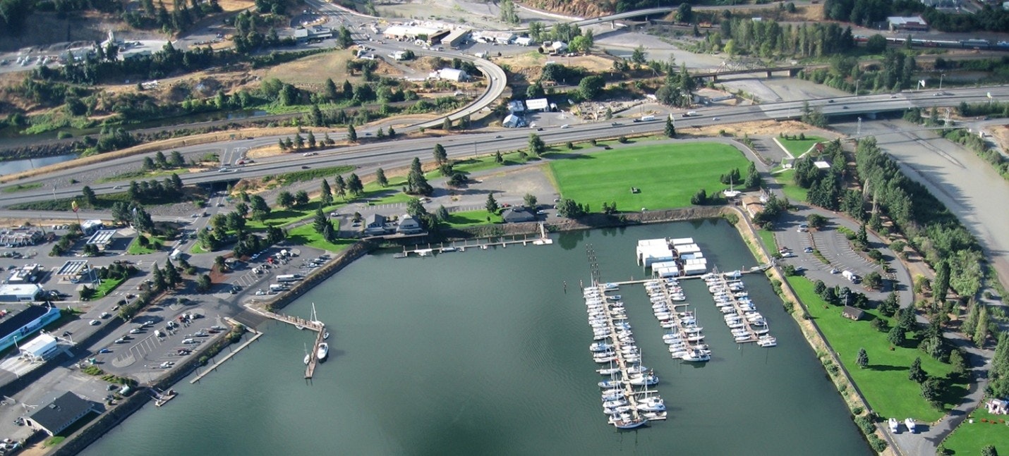 An aerial view of a marina with boats, nearby roads, and green areas, showcasing a mix of urban and natural landscapes.