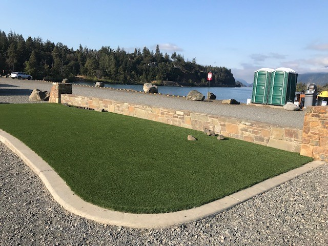 A green lawn with a stone border leading to a gravel area by the water with portable toilets and trees in the background.